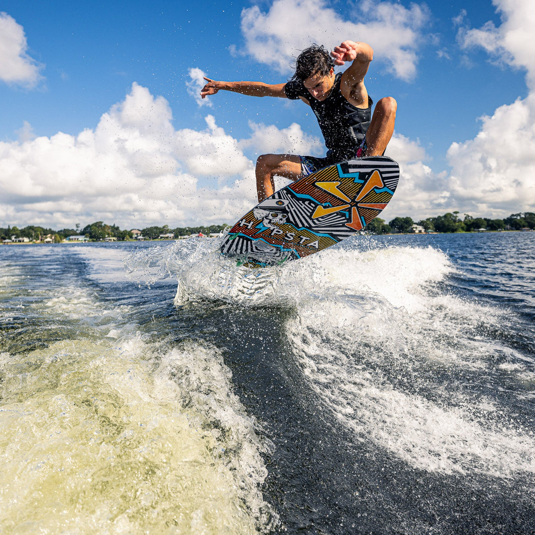 A person performs a jump on a colorful wakeboard over a wave, showcasing the 2026 Phase Five Hypsta V2 in action on a lake.