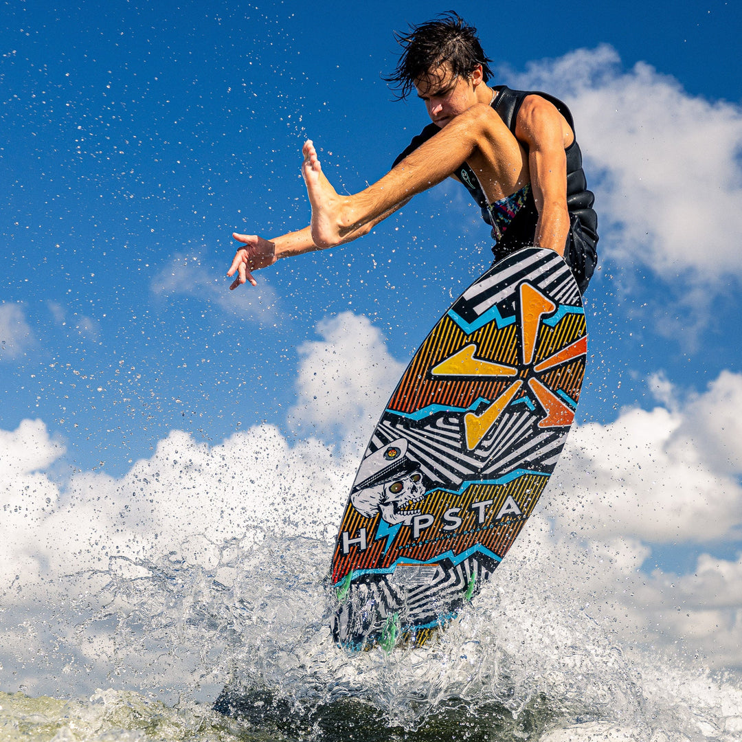 A rider performs a trick on the 2026 Phase Five Hypsta V2 wake surfboard, splashing water against a blue sky.