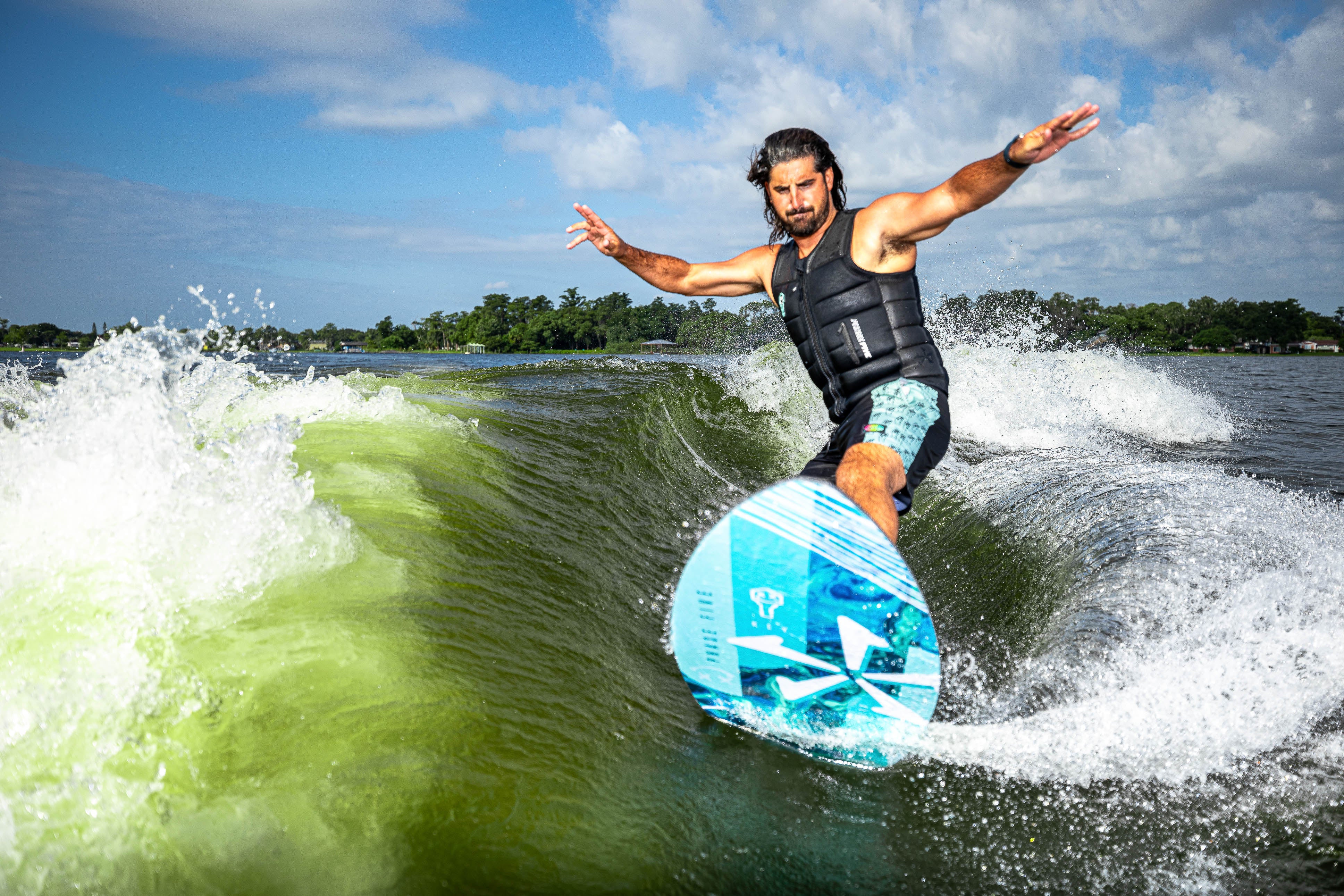 A man rides a 2025 Phase 5 Key skim board on a wave, showcasing balance and skill against a scenic water backdrop.