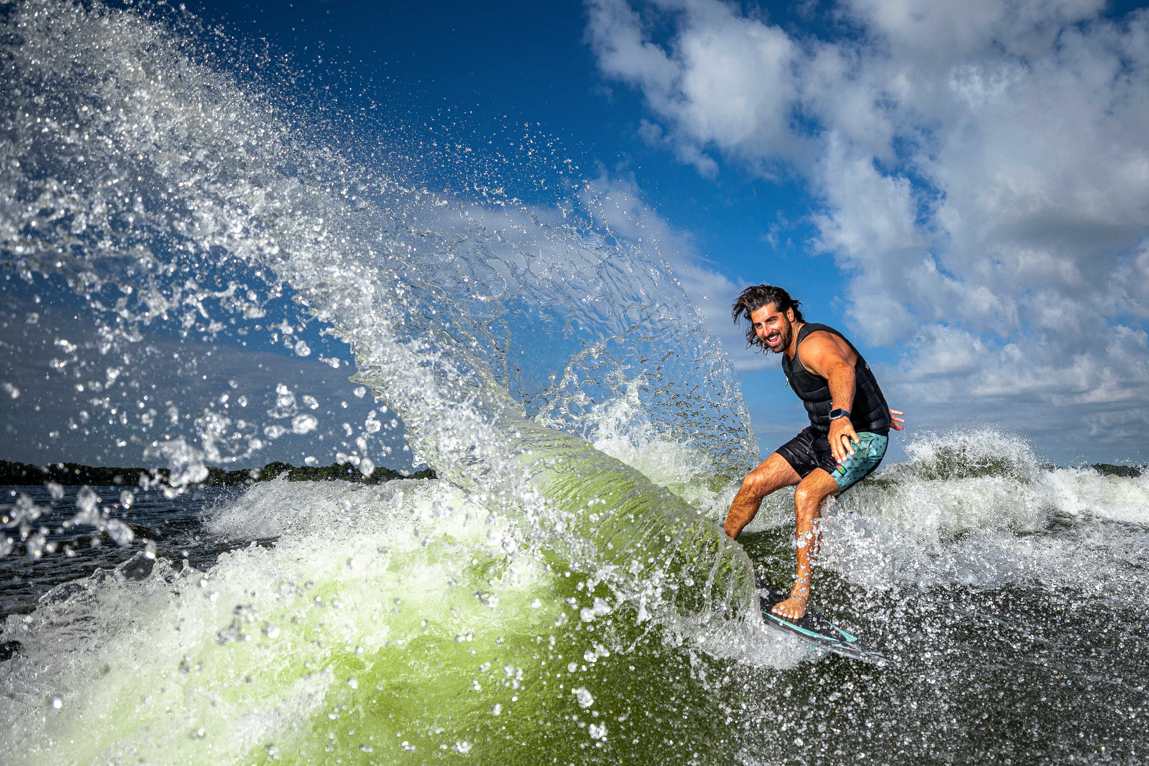 A man rides the 2025 Phase 5 Key Skim Board on a wave, showcasing skill and excitement against a vibrant sky.