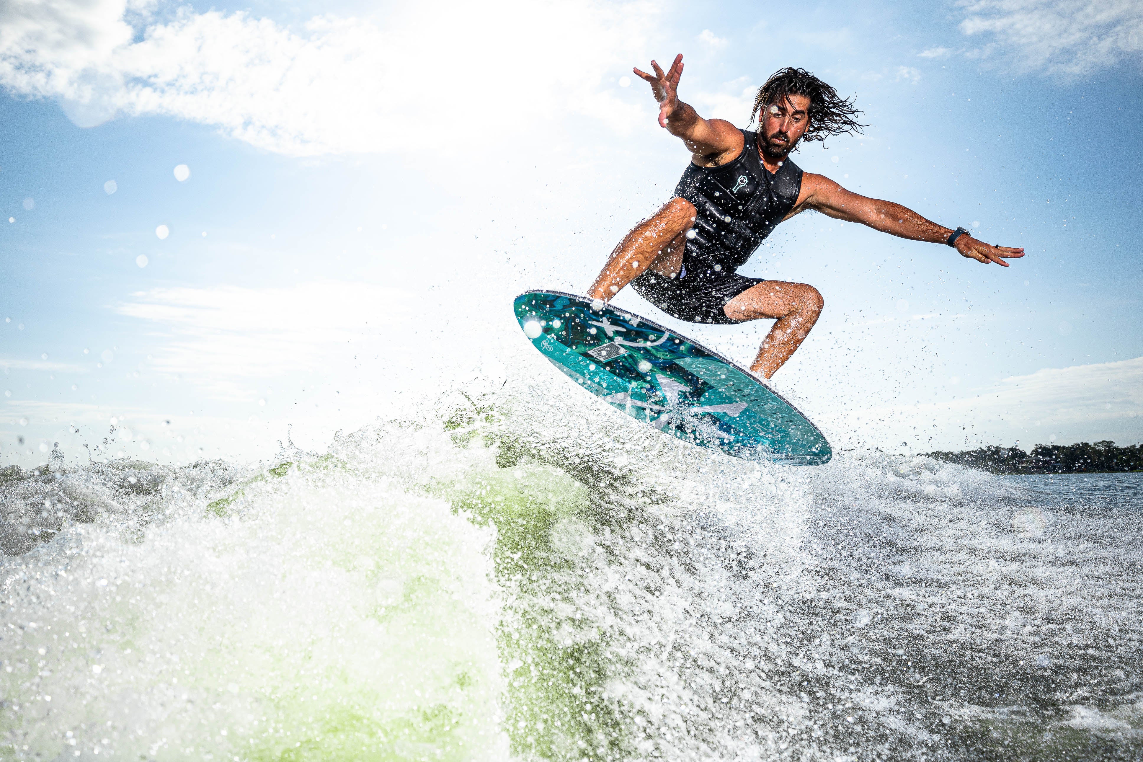A man performs a jump on a 2025 Phase 5 Key skim board over a wave, showcasing skill and excitement in water sports.