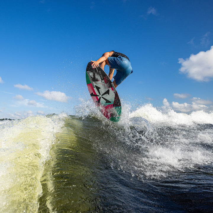 A person performing a wakeboard jump over a wave, showcasing the 2026 Phase Five Key CL board against a clear blue sky.