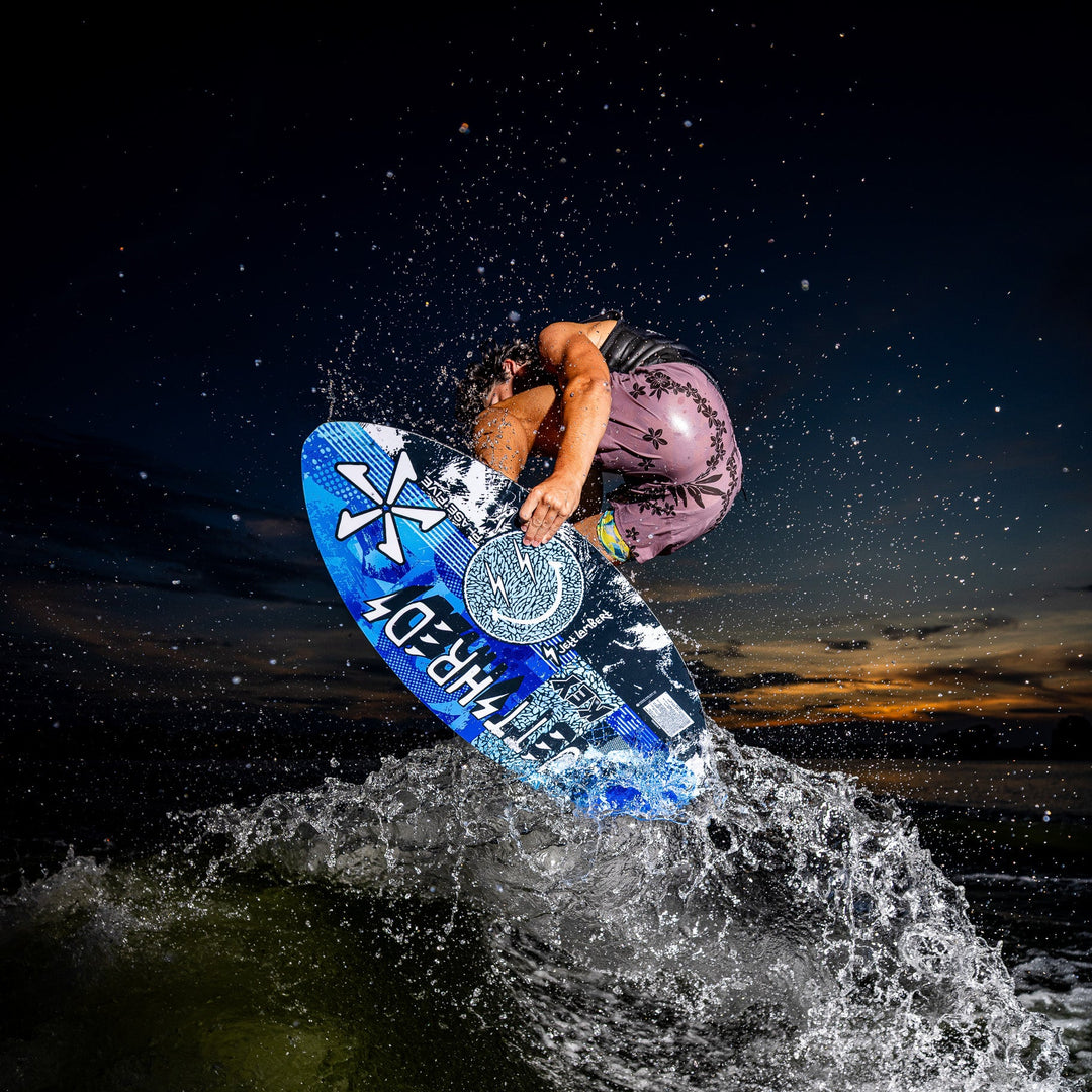 A rider performs a jump on a 2026 Phase Five Key Jett Shreds wakeboard, splashing water against a sunset backdrop.