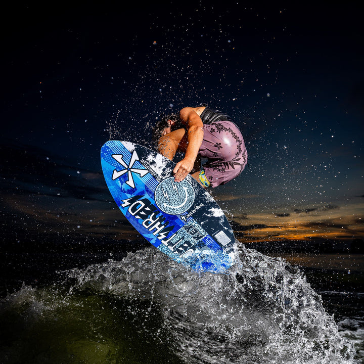 A rider performs a jump on a 2026 Phase Five Key Jett Shreds wakeboard, splashing water against a sunset backdrop.
