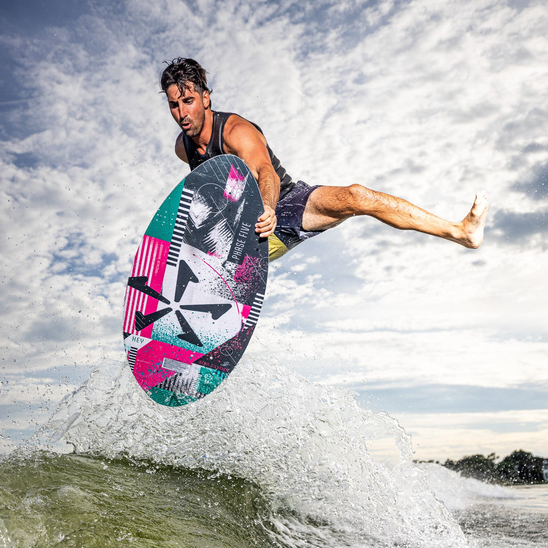A man performs a jump on a colorful Phase Five Key V2 wake surfboard above a wave, showcasing dynamic movement and skill.