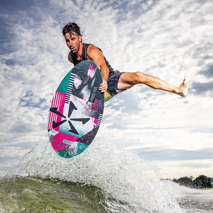A man performs a jump on a colorful Phase Five Key V2 wake surfboard above a wave, showcasing dynamic movement and skill.