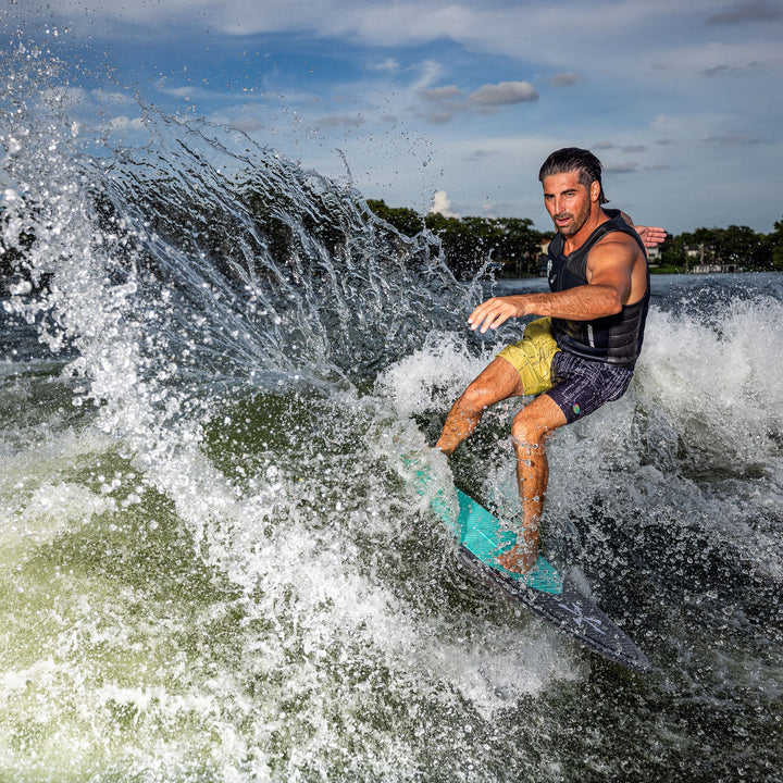 A man performs a wake surf maneuver on a 2026 Phase Five Key V2 board, creating splashes in the water.