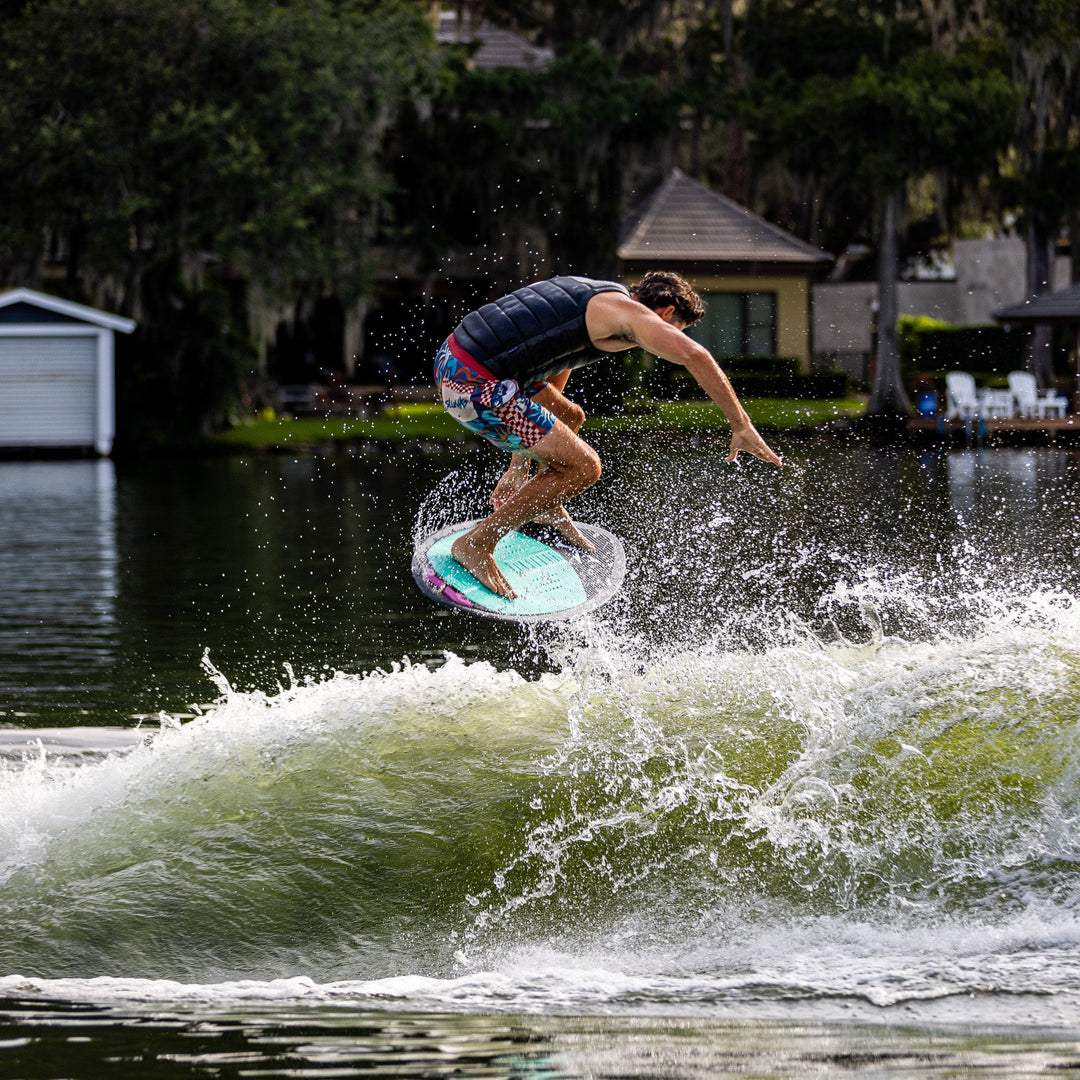 A person performs a wake surf trick on a Phase Five Key V2 board, creating splashes on a wave in a scenic lake setting.