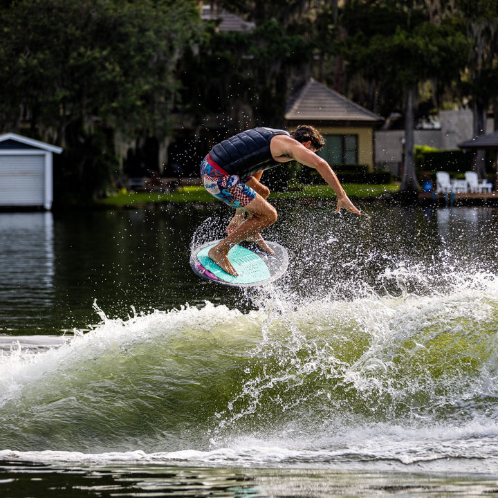 A person performs a wake surf trick on a Phase Five Key V2 board, creating splashes on a wave in a scenic lake setting.
