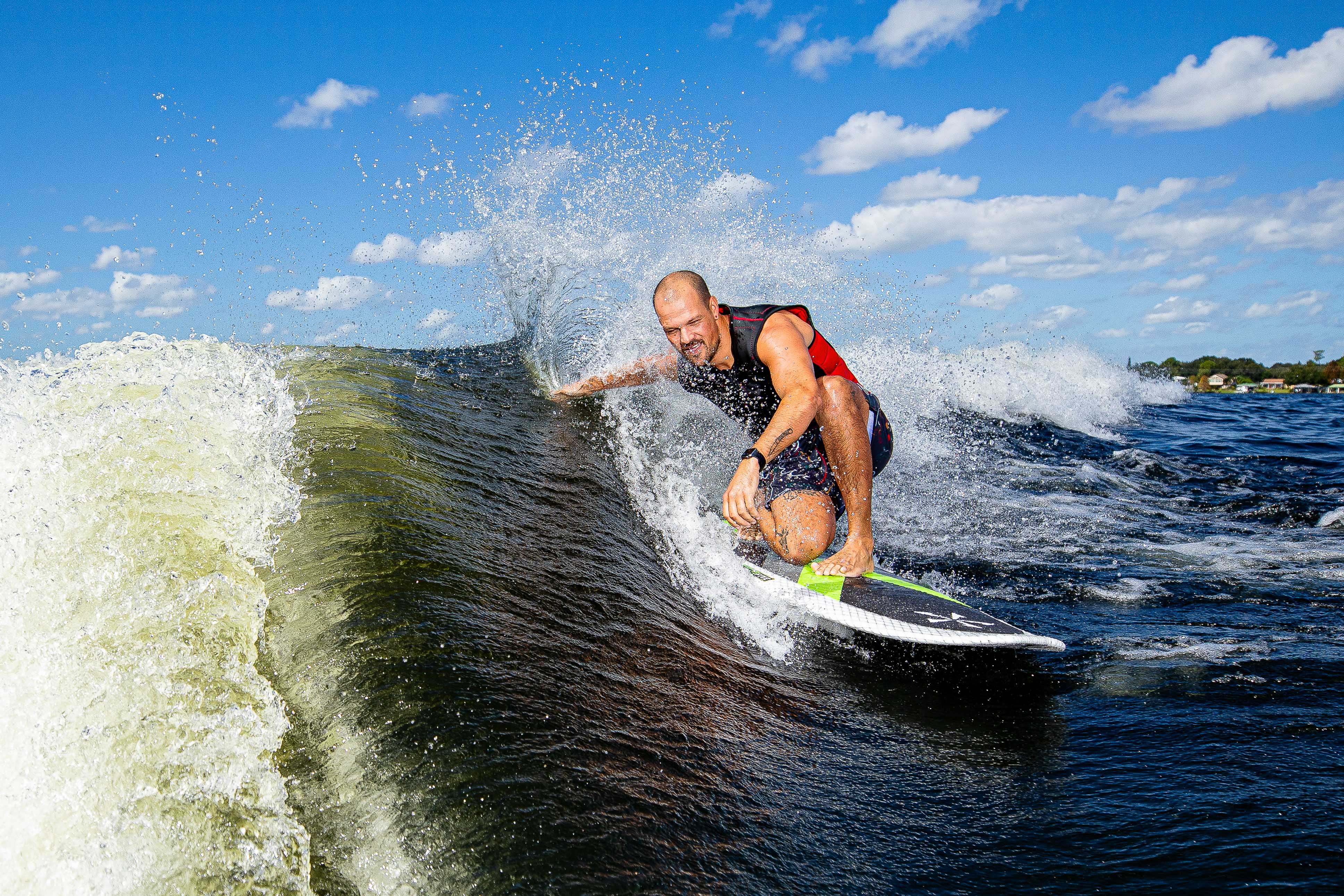 A man surfing on the 2025 Phase 5 Kong Surf Board, riding a wave with a bright blue sky and clouds in the background.