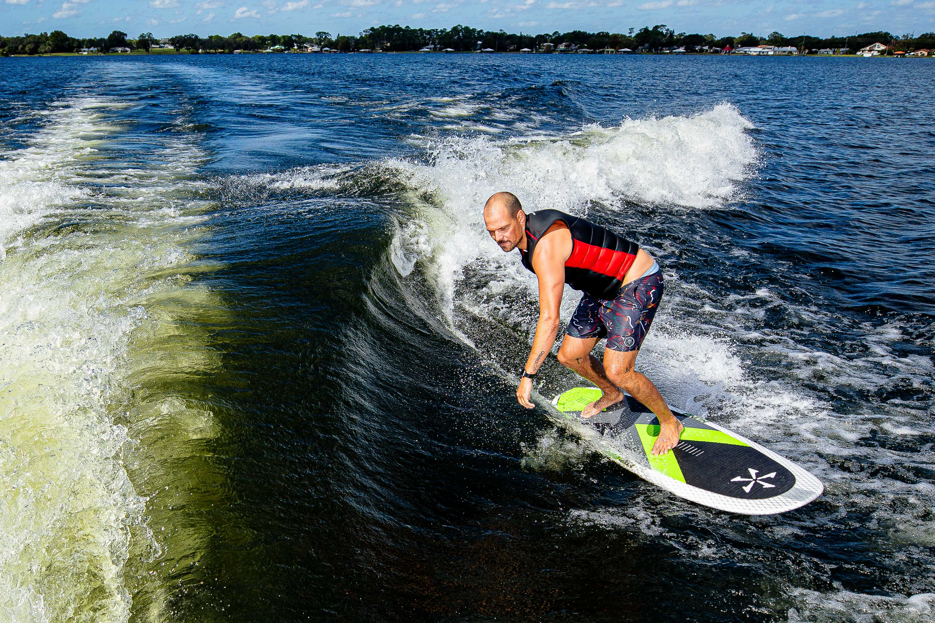 Man riding a wakeboard on a lake, wearing a life vest, with houses and trees in the background.