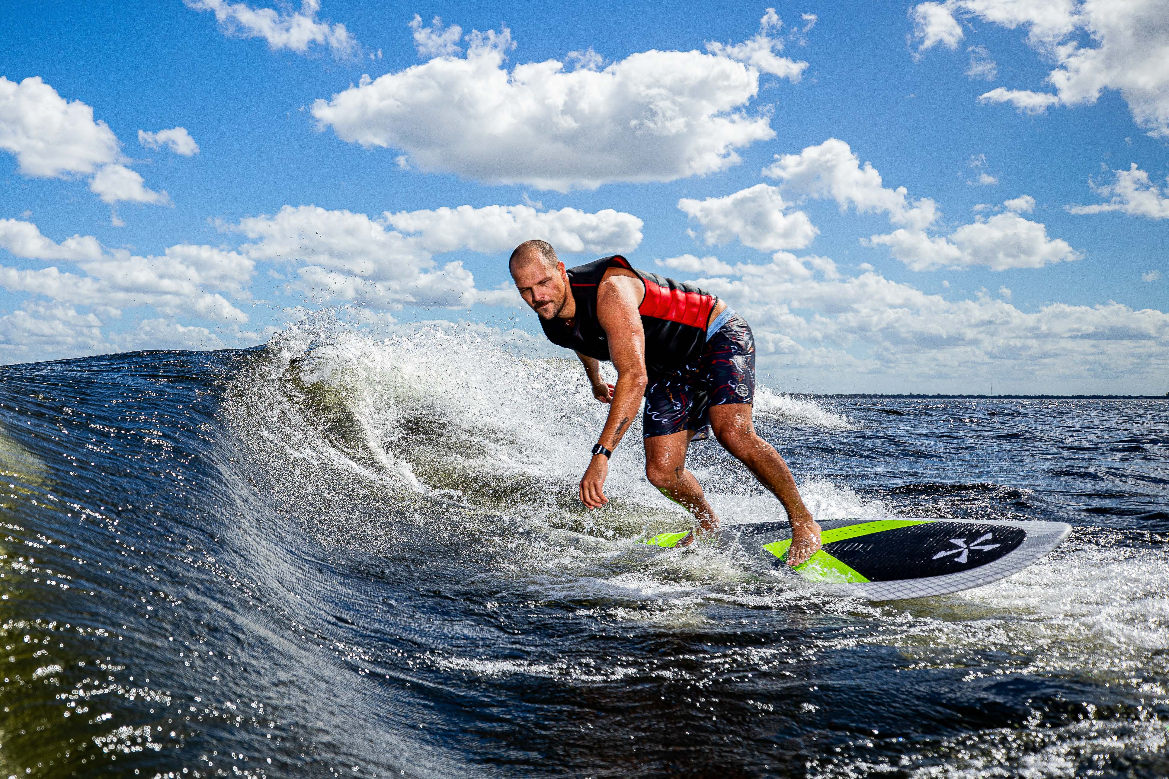 A man surfing on a green and black Kong Surf Board on a sunny day with blue skies and scattered clouds.
