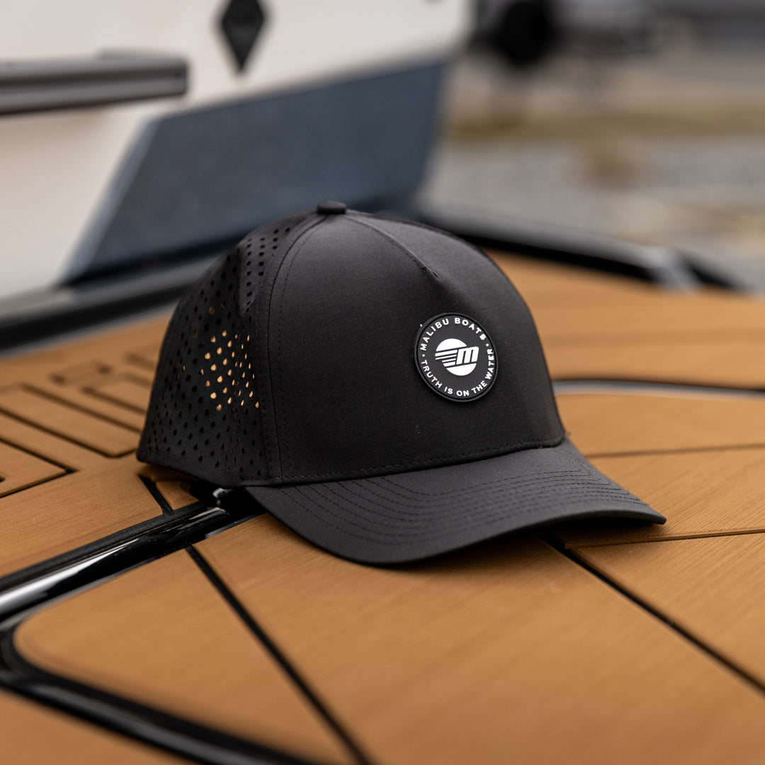 Black Malibu Truth Hat with mesh panels and a circular logo patch, resting on a wooden boat deck.