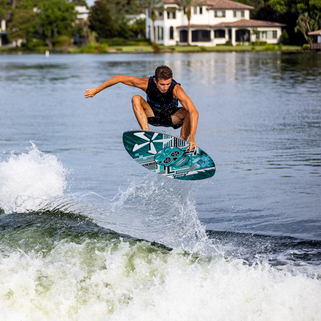 A person performs a jump on a 2026 Phase Five Matrix Pro wakeboard over a wave, with a scenic waterfront in the background.