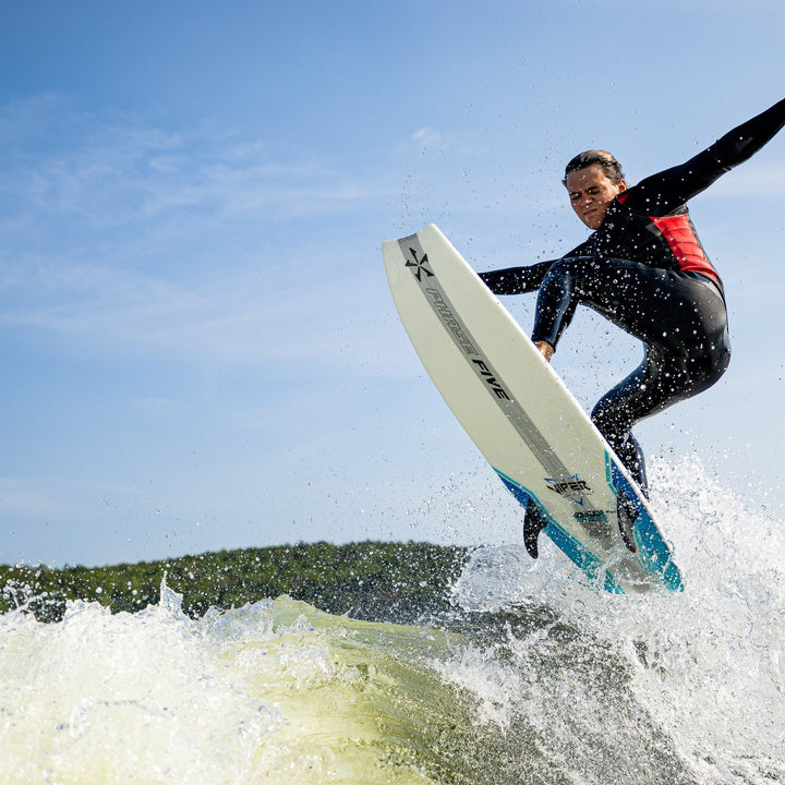 A man in a Phase Five fullsuit performs a jump on a surfboard, creating splashes in the water under a clear sky.