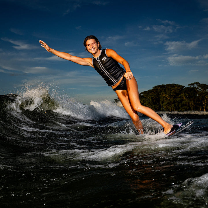 A woman surfing on a wave wearing a black life vest, showcasing the 2026 Phase Five Mindset board against a scenic backdrop.