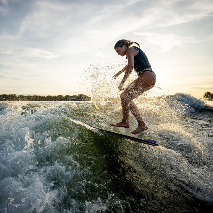 A young woman surfing on a wakeboard, creating splashes in the water during a sunset.