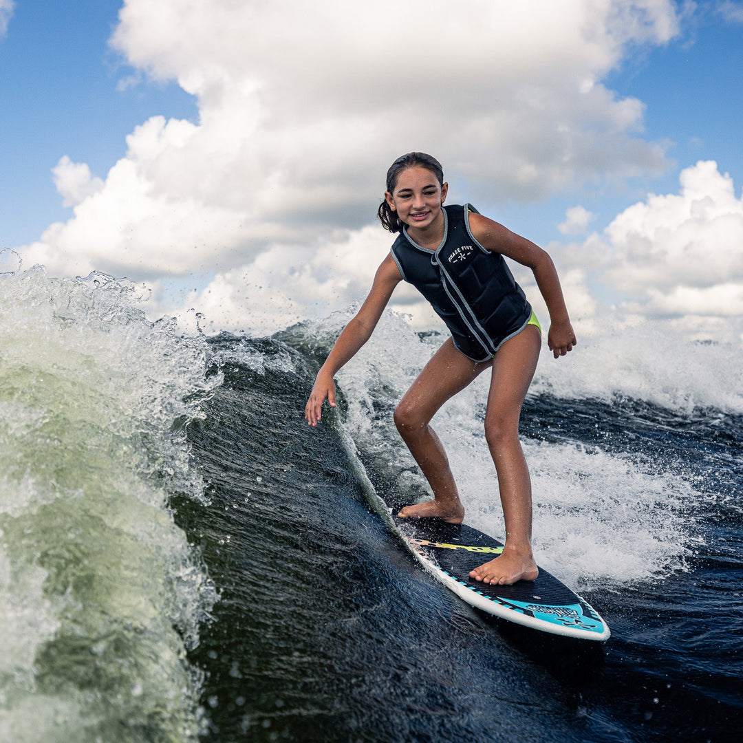 A young girl rides a wave on a 2026 Phase Five Minnow surfboard, showcasing her skills in a life jacket.