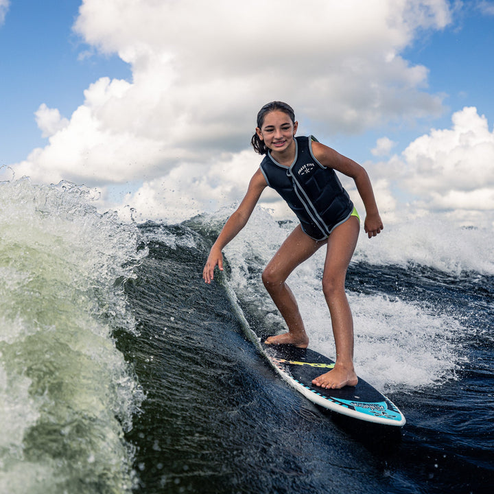 A young girl rides a wave on a 2026 Phase Five Minnow surfboard, showcasing her skills in a life jacket.