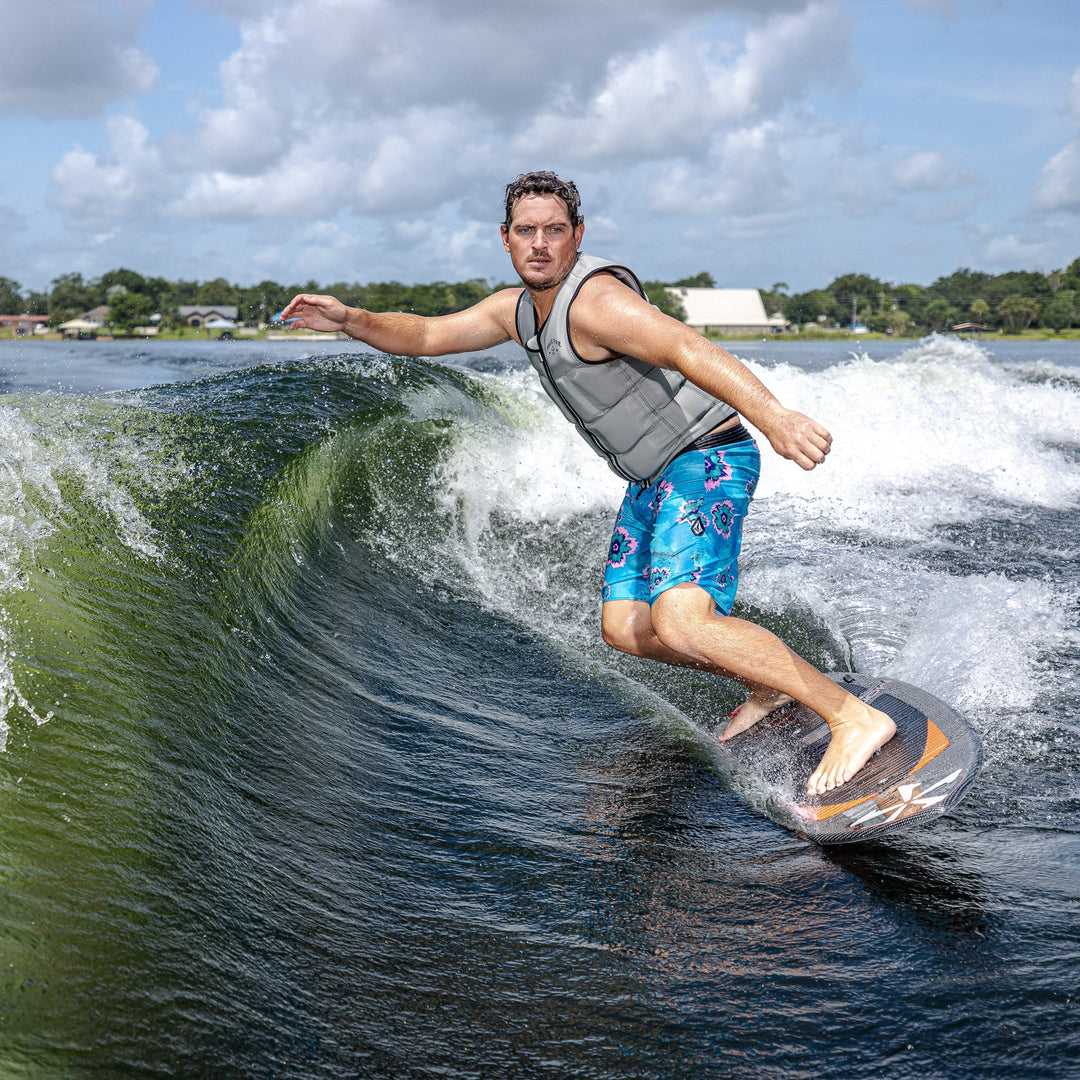 A man surfing on a 2026 Phase Five Model X wakesurf board, riding a wave with a scenic background.