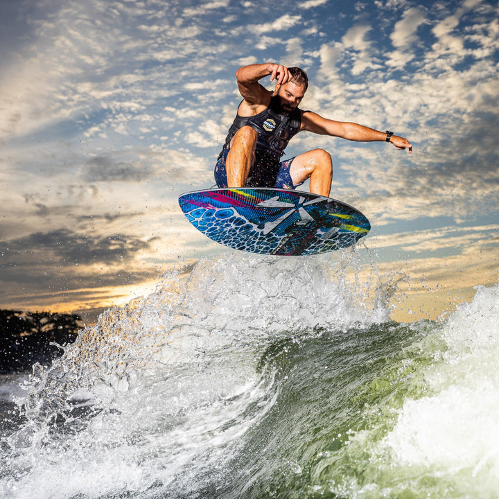 A man performs a jump on a wakeboard over a wave during sunset, showcasing the 2026 Phase Five MVP V4 design.