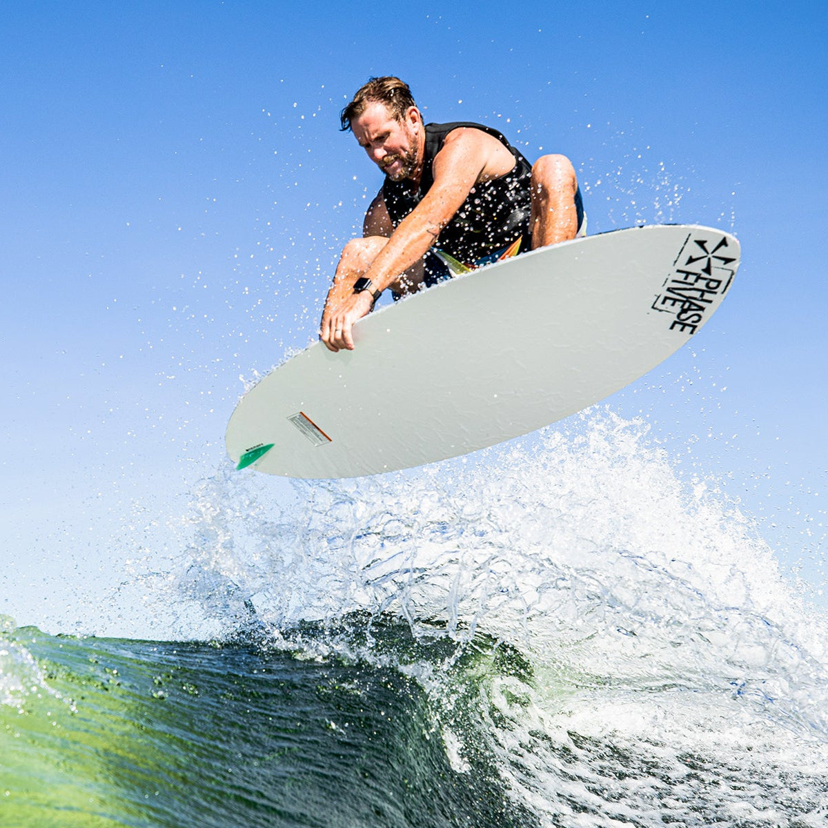 A man performs an aerial maneuver on a 2025 Phase 5 Oogle skim board above a wave.
