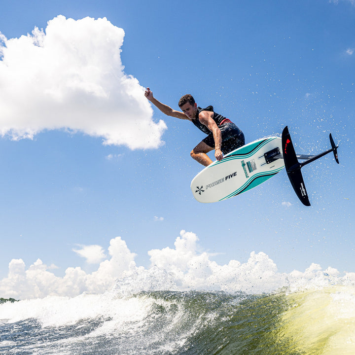 A rider performs a jump on the 2026 Phase Five Phantom Foil above ocean waves under a clear blue sky.