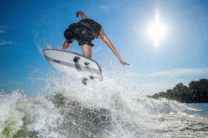 A person performing an aerial trick on a white Phantom Stealth Surf Board during daytime with a bright sun and blue sky.