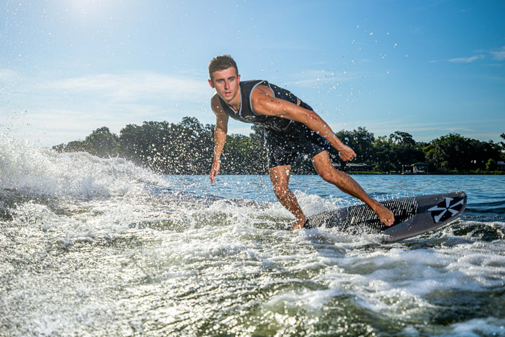 A young man surfing on the 2025 Phase 5 Phantom Stealth Surf Board on a lake, with water splashing and a clear blue sky.