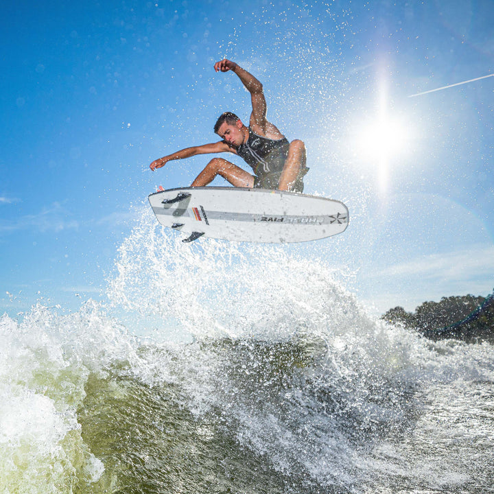 2025 Phase 5 Phantom Stealth Surf Board being used by a surfer jumping over a wave under a bright sun.