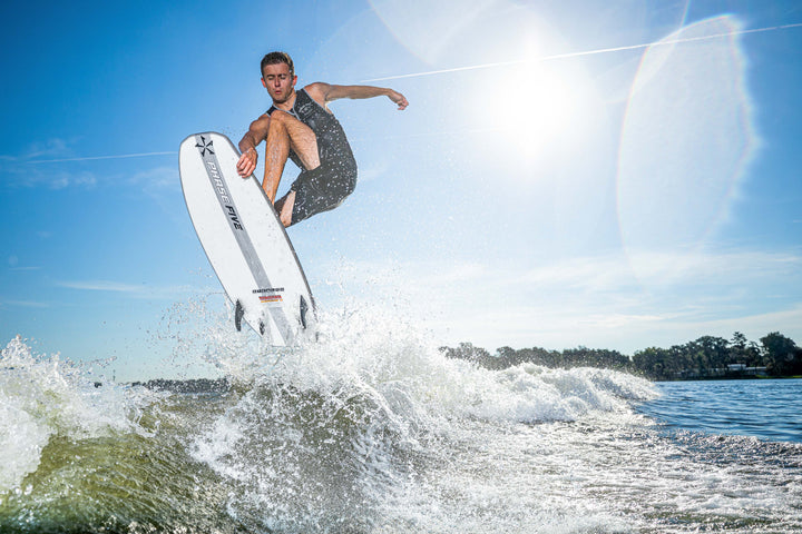 A man performs an aerial trick on a white Phantom Stealth Surf Board during sunny daytime water sports activity.