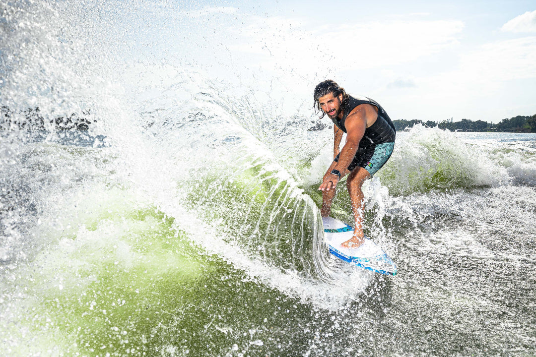 A person rides the 2025 Phase 5 Prop Skim Board on a wave, showcasing dynamic movement and water spray.