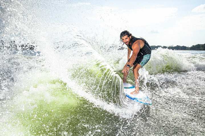 A person rides the 2025 Phase 5 Prop Skim Board on a wave, showcasing dynamic movement and water spray.