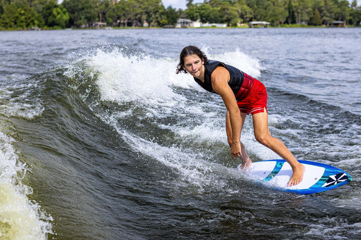 A person rides a 2025 Phase 5 Prop Skim Board on a wave, showcasing dynamic movement and water spray.