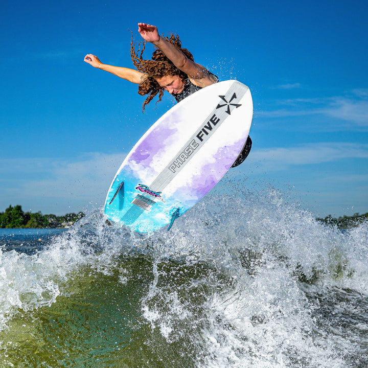 A female surfer performing an aerial trick on a colorful Phase 5 Rush surfboard, riding a wave under a clear blue sky.