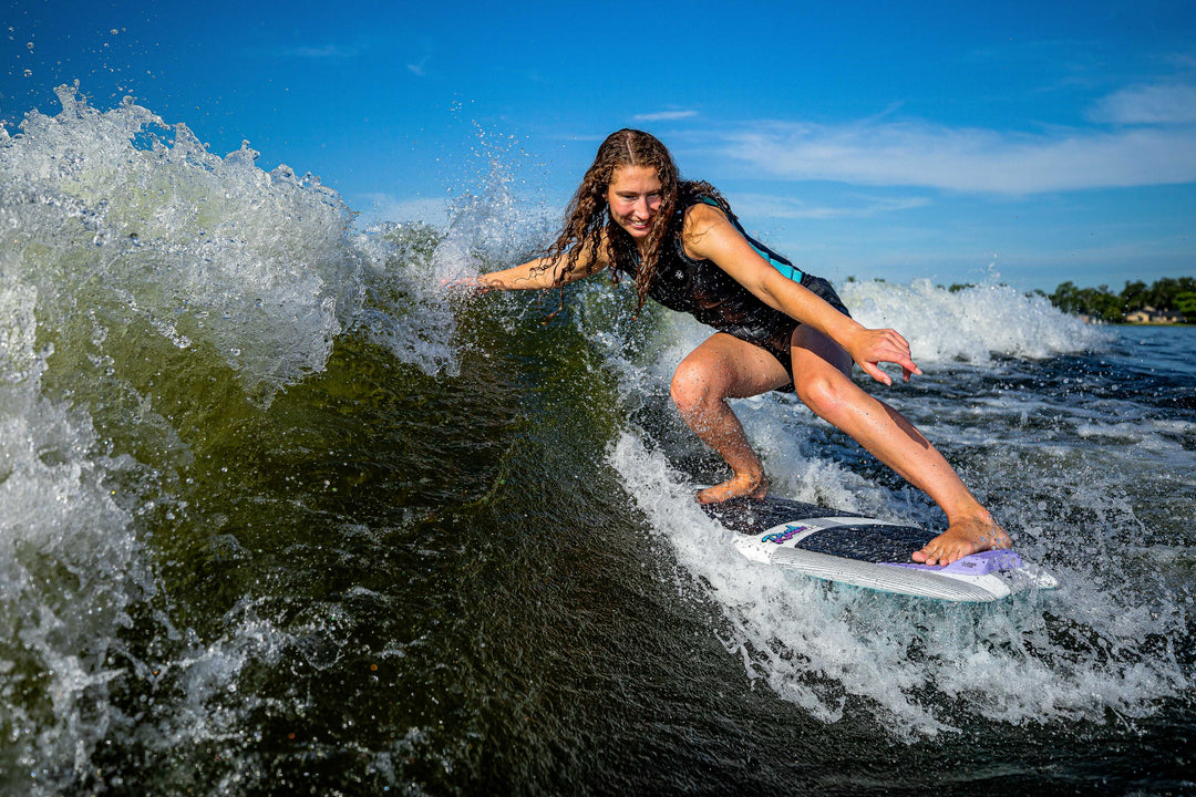 A woman surfing on a 2025 Phase 5 Rush surfboard, riding a wave with a bright blue sky in the background.