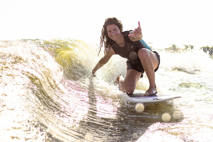 Female surfer riding a wave on the 2025 Phase 5 Rush surfboard, smiling and making an "L" gesture with her hand.