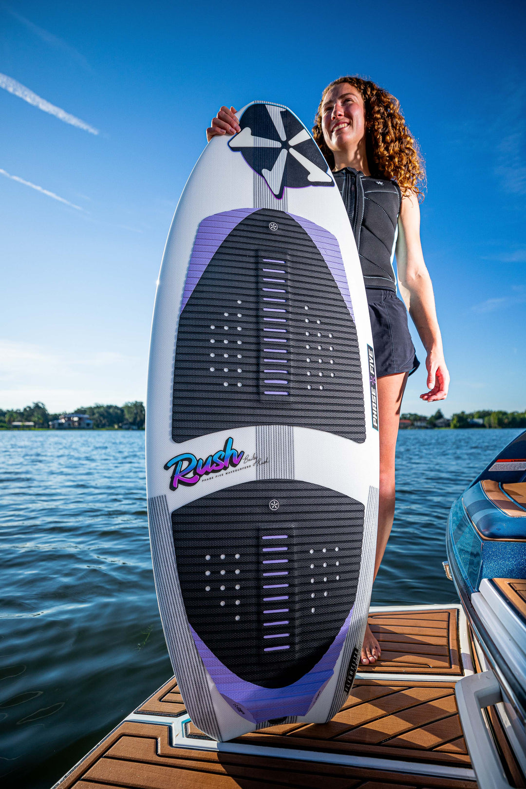 A woman holding a 2025 Phase 5 Rush Surf Board on a boat dock, with water and a clear blue sky in the background.