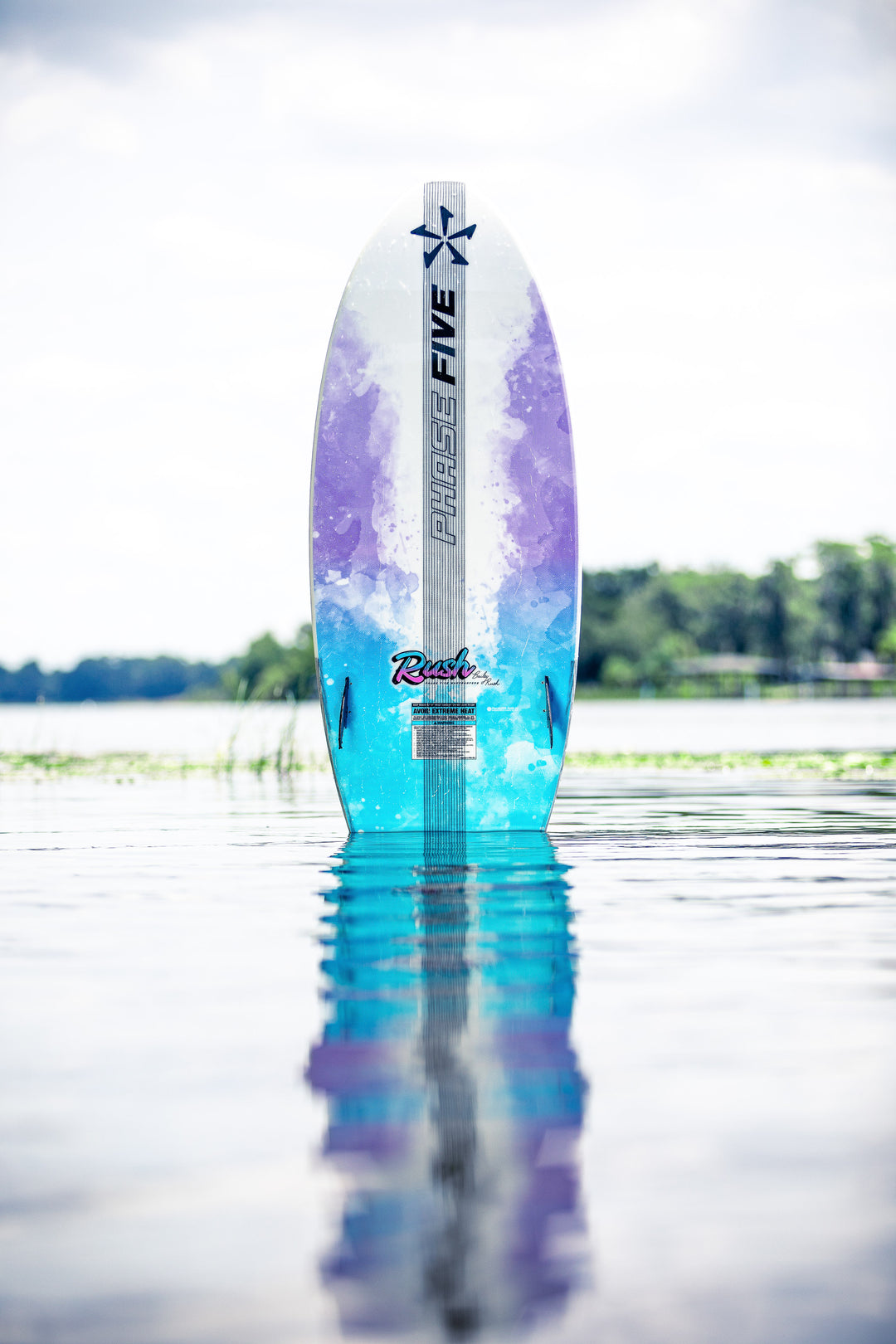 2025 Phase 5 Rush surfboard with colorful purple, blue, and white design, standing upright in calm water near a lakeshore.
