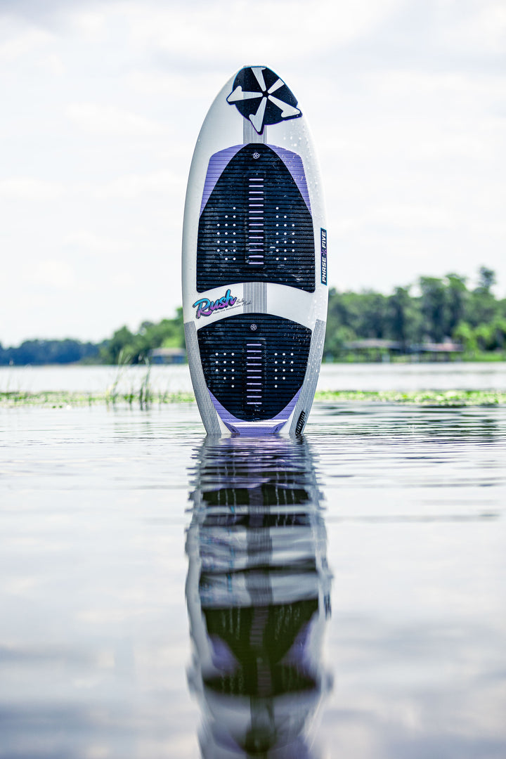 2025 Phase 5 Rush surfboard standing upright in calm water with a scenic background and reflection on the surface.