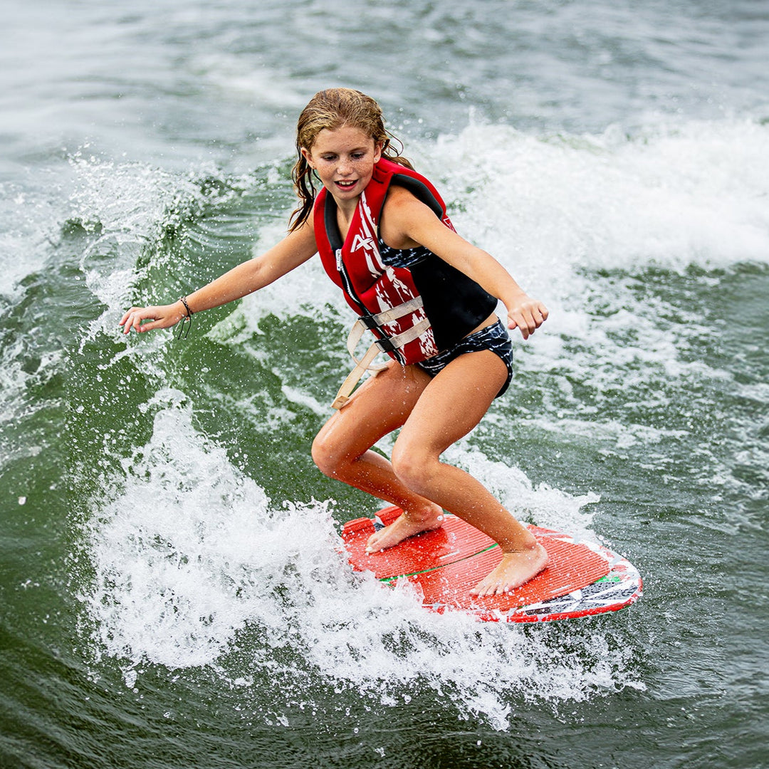 A young girl rides a 2025 Phase 5 Shrimp Skim Board on a wave, showcasing her skills in water sports.