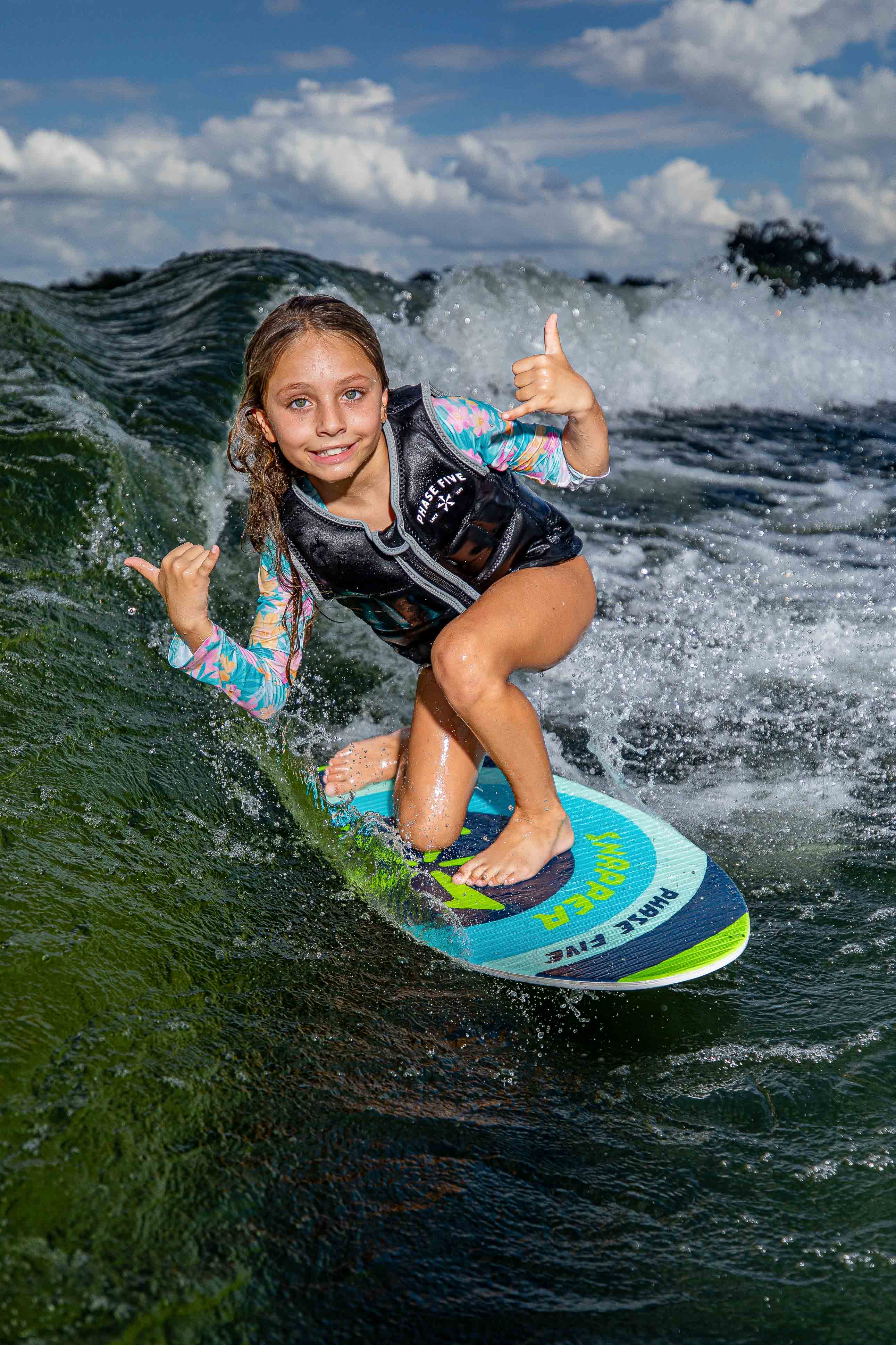 Young girl riding a Snapper Skim Board on water, wearing a life vest, with a blue sky and clouds in the background.