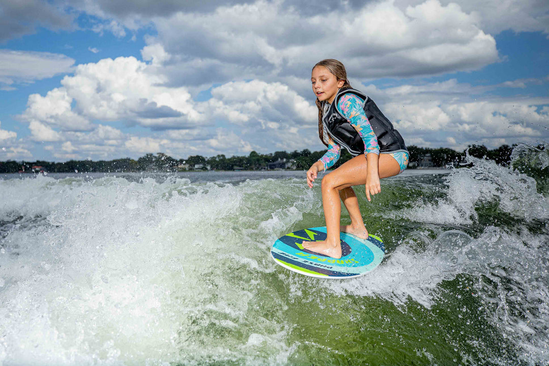Young girl surfing on a blue and green Snapper Skim Board in the water under a partly cloudy sky.