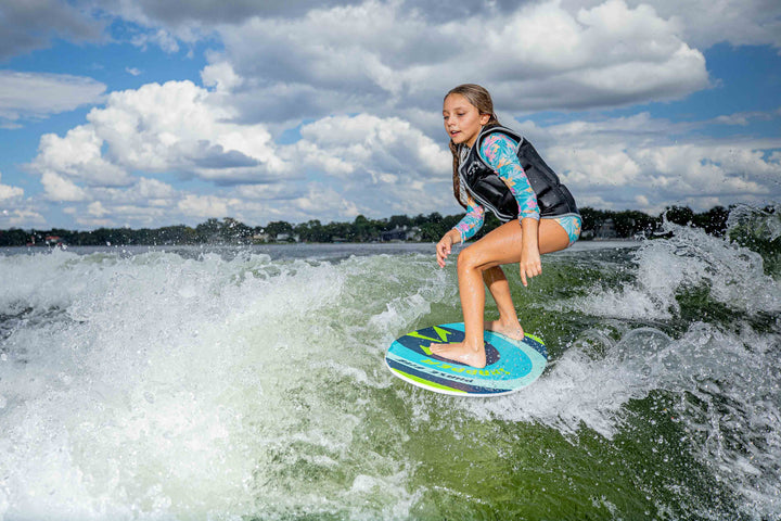 Young girl surfing on a blue and green Snapper Skim Board in the water under a partly cloudy sky.