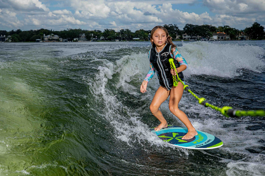 A girl surfing on a blue and green Snapper Skim Board on a lake, wearing a life vest and colorful long-sleeve shirt.