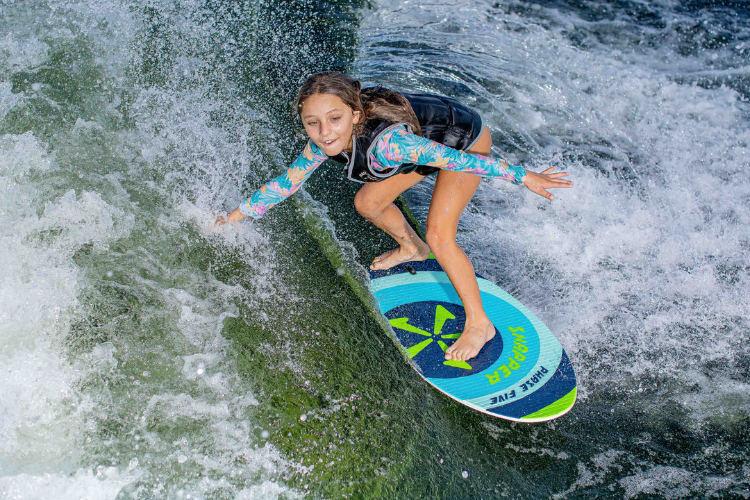 Young girl surfing on a blue and green Snapper Skim Board in the water, wearing a colorful long-sleeve top and black vest.
