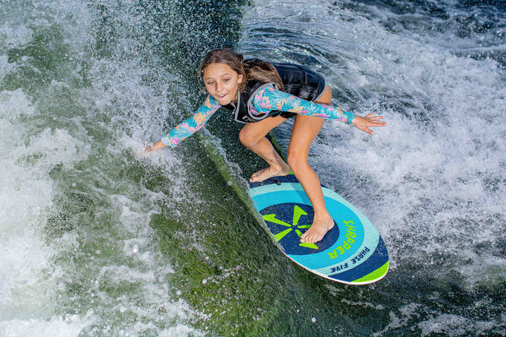 Young girl surfing on a blue and green Snapper Skim Board in the water, wearing a colorful long-sleeve top and black vest.