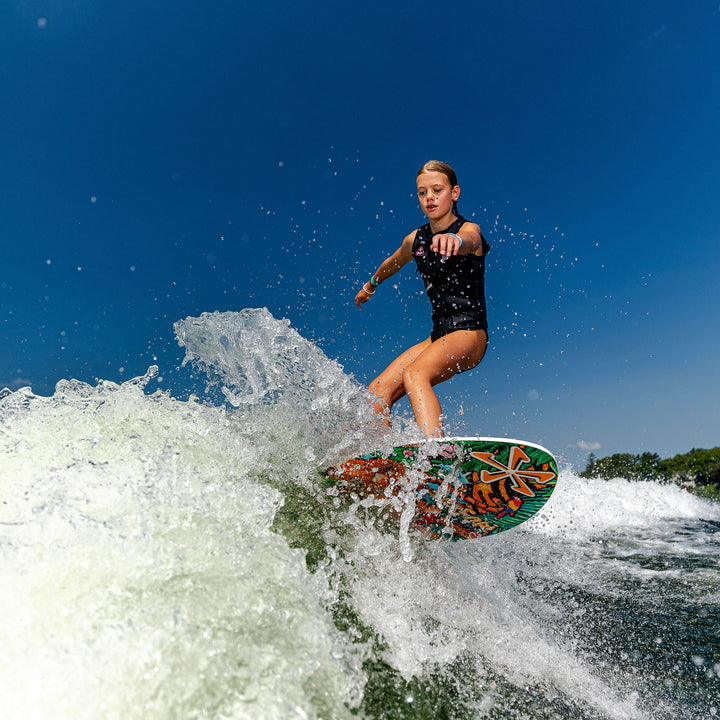 A young girl surfing on the 2026 Phase Five Surfari board, riding a wave under a clear blue sky.
