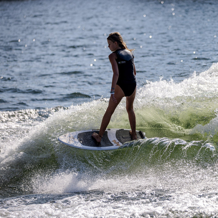 A young girl rides the 2026 Phase Five Surfari on a wave, showcasing balance and skill in a vibrant water setting.