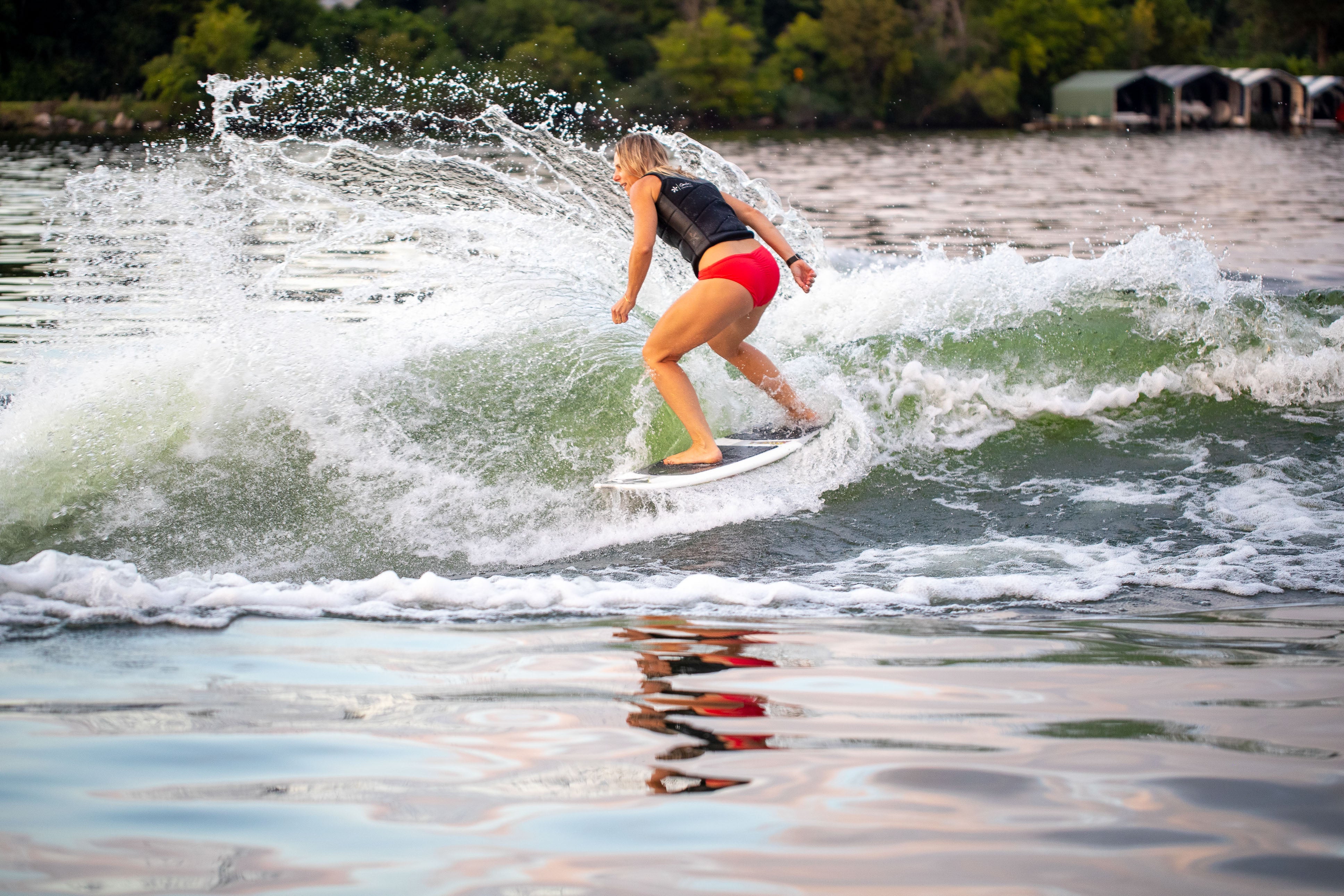 A woman surfing on a 2025 Phase 5 Swell Surf Board, riding a wave near a lakeshore with trees and boats in the background.
